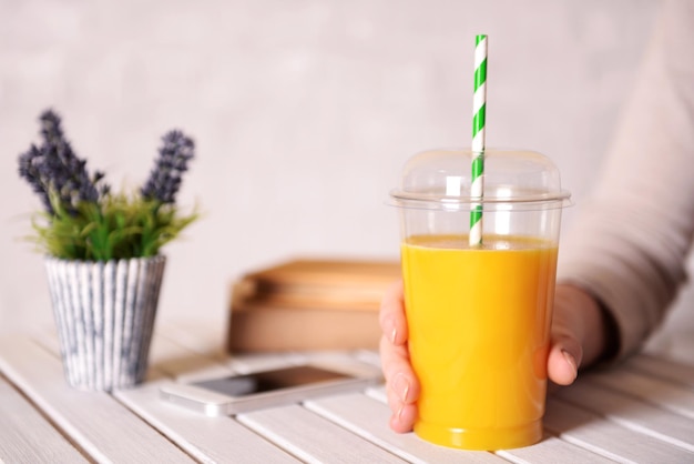 Vrouwelijke hand aan houten tafel met fastfood gesloten kopje sinaasappelsap en in de buurt van boekenplant en mobiele telefoon op lichte muurachtergrond