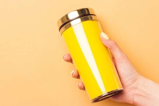 Vrouwelijke hand die een gele lege thermocup voor dranken op een warme gele achtergrond houdt