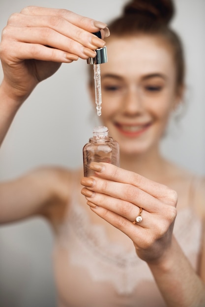 Foto vrouwelijke hand die een hyaluronzuurfles vasthoudt en de druppelaar tegen een witte muur neemt.