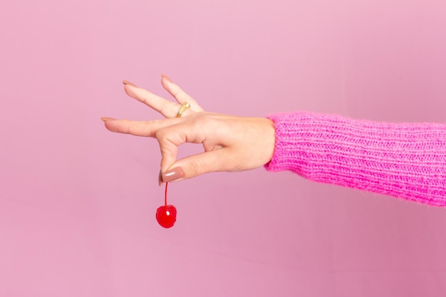 Foto vrouwelijke hand die roze sweater draagt en een kers houdt. licht roze achtergrond.