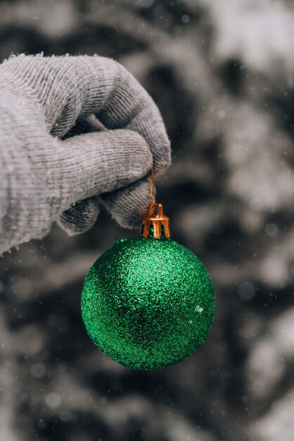 Foto vrouwelijke hand in handschoenen groene glanzende trendy kerstbal op besneeuwde taksparren in de winter bosvakantie