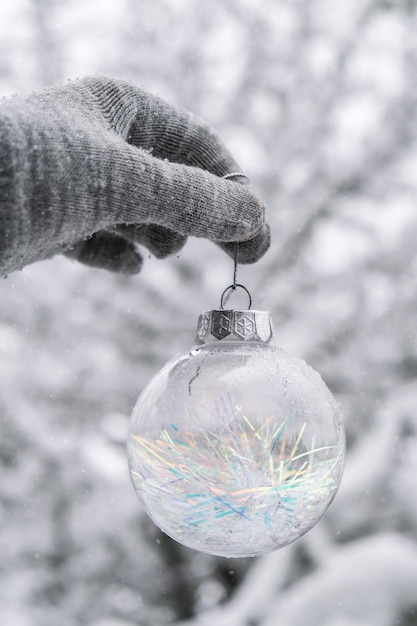 Foto vrouwelijke hand in handschoenen transparant trendy glazen kerstbal op besneeuwde taksparren in winterbos