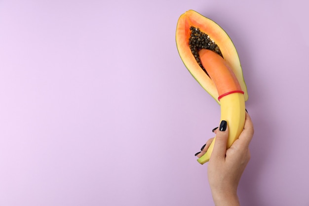 Vrouwelijke hand met banaan met condoom in papaya op violette achtergrond