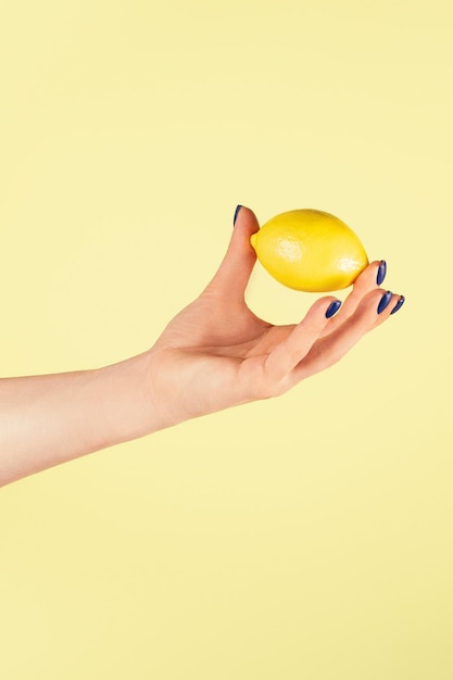 Vrouwelijke hand met blauwe spijkers die lichtjes met twee vingers verse rijpe sappige citroen houden die op geel wordt geïsoleerd. Creatief kunstconcept. Voedsel minimaal kleurrijk concept. Verticaal. Ruimte kopiëren. Bespotten.