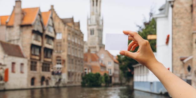 Vrouwelijke hand met een kaartje of visitekaartje met kopieerruimte op de achtergrond van een gracht en middeleeuwse huizen in Brugge, België