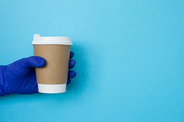 vrouwelijke hand met een kopje koffie kopje. Kopieer ruimte. Koffiekop ter beschikking met medische handschoenen die op blauwe achtergrond worden geïsoleerd. Hand met papieren beker, coronavirusbescherming