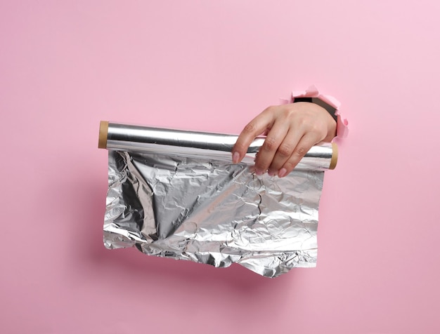 Vrouwelijke hand met een rol grijze voedselfolie op een roze achtergrond, een deel van het lichaam steekt uit een gescheurd gat
