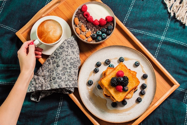 Vrouwelijke hand met yoghurtkom en bessen, wentelteefjes, koffiekopje op plaat tijdens het ontbijt op bed