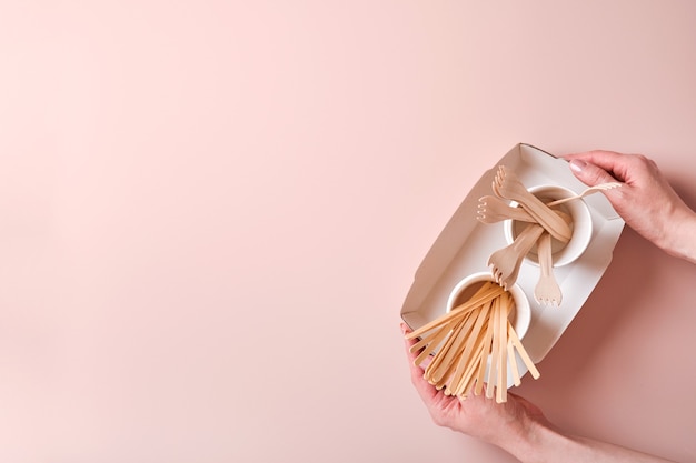 Vrouwelijke hand neemt cocktailrietjes, papieren bekers, borden, houten vorken, rietjes, fastfoodcontainers, houten bestek op roze achtergrond. eco ambachtelijk papier servies. recyclingconcept. bespotten.