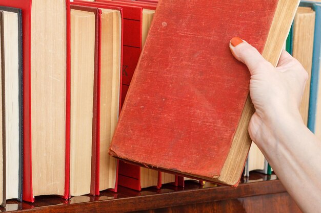 Vrouwelijke hand neemt een boek van een boekenplank. Veel boeken met harde kaft op houten plank. Bibliotheekconcept