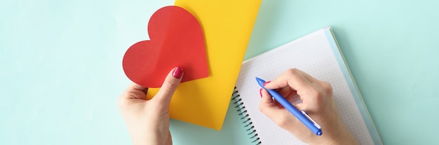 Vrouwelijke hand schrijft brief in een notitieboekje en houdt een valentijnskaart vast met een envelop in haar handen