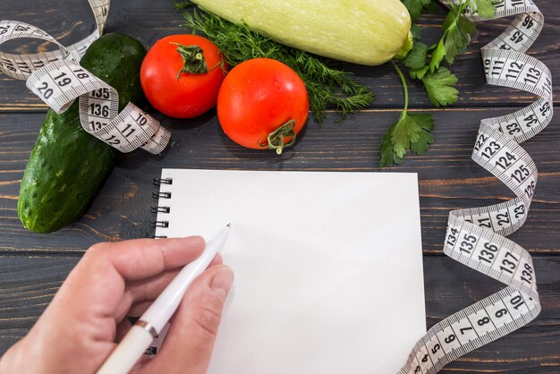 Foto vrouwelijke hand schrijft in een leeg notitieboekje. diverse groenten, meetlint en notitieblok