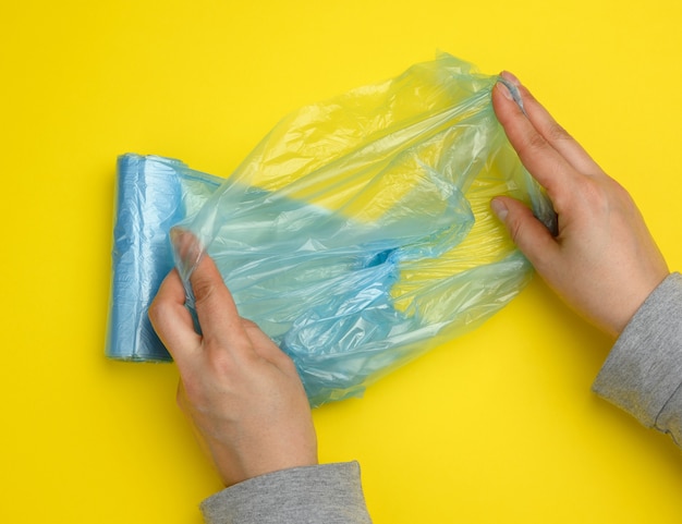 Vrouwelijke hand wikkelt een blauwe plastic zak af voor afval, gele achtergrond, bovenaanzicht