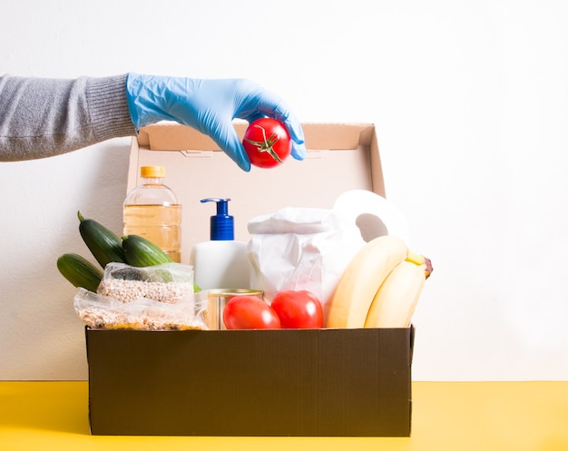 Vrouwelijke hand zet een tomaat in een doos in boodschappen, producten en hygiëneproducten voor donatie, kopie ruimte, wit en geel oppervlak