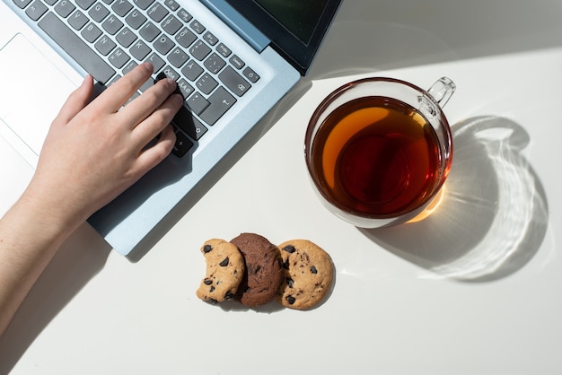 Vrouwelijke handafdrukken op een laptop, een kopje thee en drie chocoladeschilferkoekjes in zonlicht, bovenaanzicht.