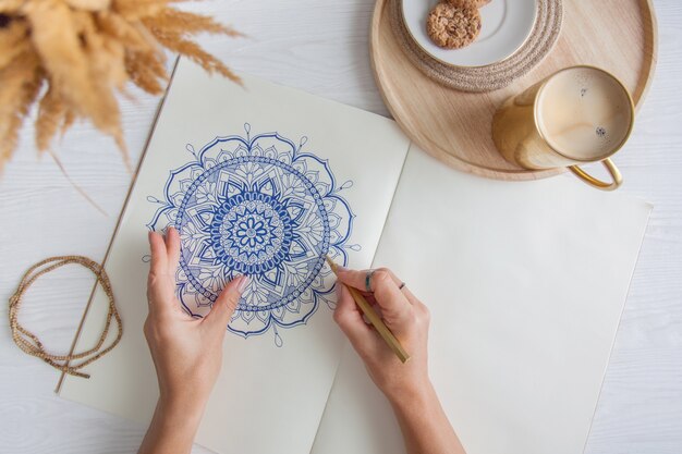 Vrouwelijke handen close-up tekenen decoratieve ronde bloemen mandala. Hobby en thuisontspanning. Een mok koffie en koekjes op een houten dienblad. Witte achtergrond.