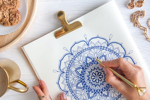 Vrouwelijke handen close-up tekenen decoratieve ronde bloemen mandala. Hobby en thuisontspanning. Een mok koffie en koekjes op een houten dienblad. Witte achtergrond.