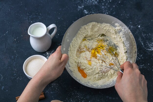 Vrouwelijke handen die deeg voorbereiden en bloem en eieren zwaaien. Ingrediënten en keukengerei voor het koken van cake (meel, ei, melk, suiker, deegroller, handdoek) op donkere tafel. Het concept van het maken van deeg voor het bakken