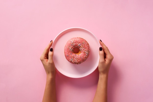 Vrouwelijke handen die doughnut op plaat over roze achtergrond houden.