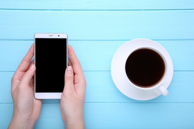 Vrouwelijke handen die smartphone en kop van koffie op blauwe houten achtergrond houden.