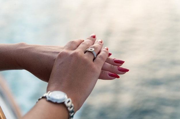 Vrouwelijke handen en polshorloge met rode manicure en gouden ring aan de vinger met diamanten in de buurt van water