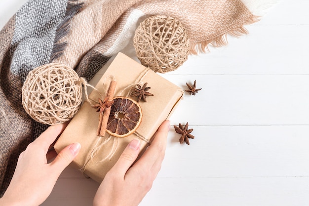 Foto vrouwelijke handen houden een geschenkdoos in ambachtelijk papier met anijs, droge kegels, sinaasappel en kaneelstokje op een witte houten in rustieke stijl, copyspace, bovenaanzicht