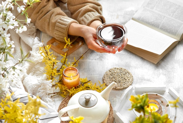 Vrouwelijke handen houden een kopje thee tegen een tafel met gezellige decordetails en gele bloemen.