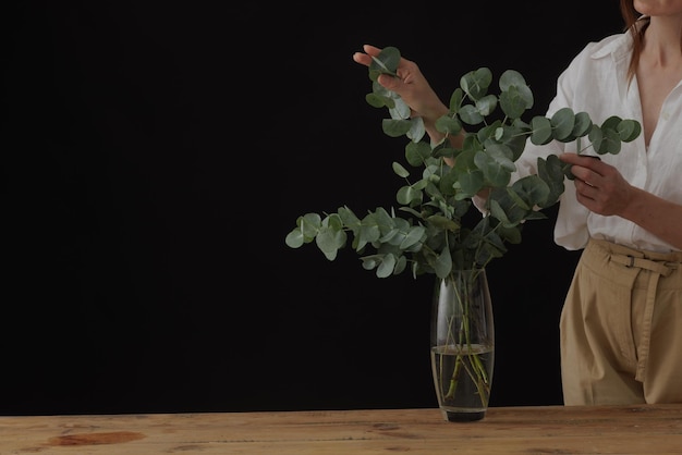 Vrouwelijke handen houden eucalyptus in glazen vaas op houten tafel donkere achtergrond mockup