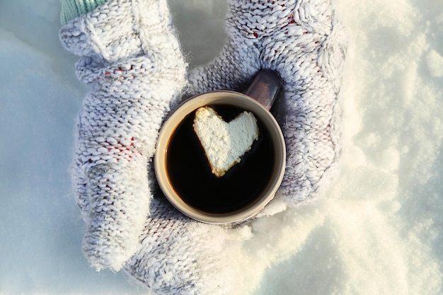 Vrouwelijke handen in warme wanten met kop warme koffie met hart marshmallow op besneeuwde achtergrond