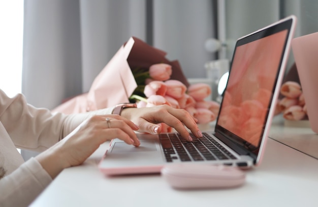Vrouwelijke handen met behulp van laptop. Vrouwelijk bureau werkruimte thuiskantoor mock-up met laptop, roze tulp bloemen boeket, smartphone en roze accessoires.