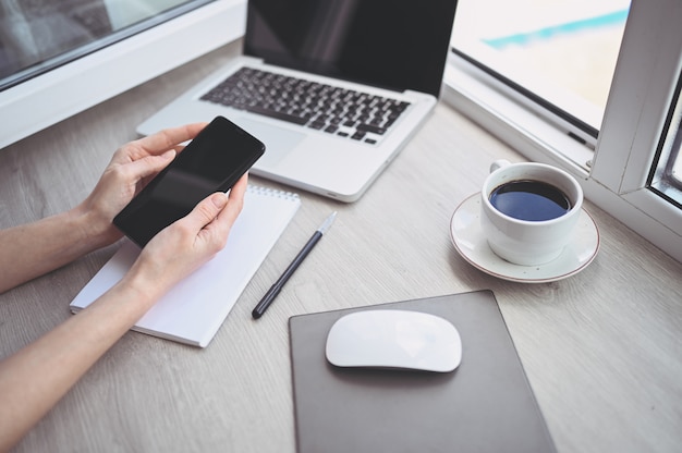 Vrouwelijke handen met behulp van slimme telefoon in interieur werkplek, freelance zakenvrouw met behulp van mobiele telefoon op kantoor, werken vanuit huis met behulp van slimme telefoon en notebook computer. Quarantaine op afstand