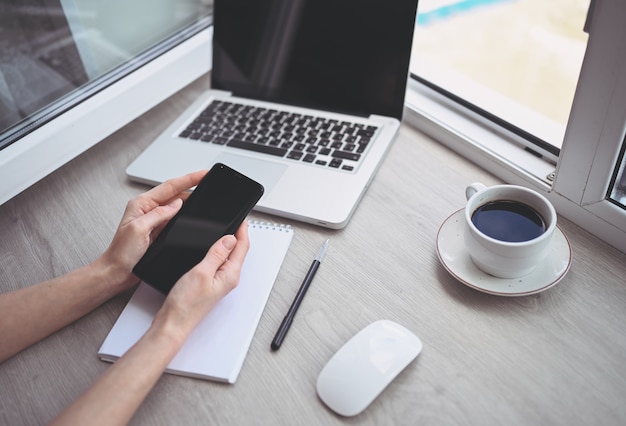 Vrouwelijke handen met behulp van slimme telefoon in interieur werkplek, freelance zakenvrouw met behulp van mobiele telefoon op kantoor, werken vanuit huis met behulp van slimme telefoon en notebook computer. Quarantaine op afstand