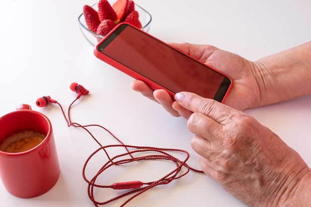 Vrouwelijke handen met behulp van telefoon met rood scherm. witte desktop met koffiekopje en aardbeien