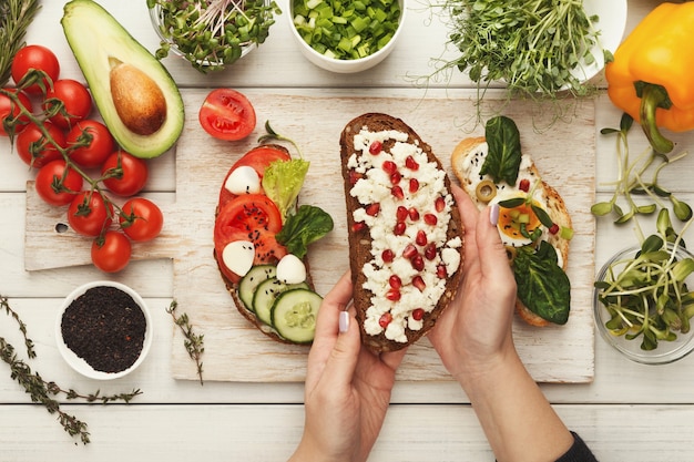 Vrouwelijke handen met bruschetta met fetakaas en granaatappelzaden. Gezonde vegetarische sandwiches aan keukentafel met diverse groentenkommen en greens. Koken voedsel achtergrond, bovenaanzicht