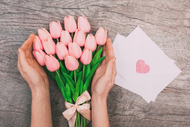 Vrouwelijke handen met een boeket van roze tulpen en lege witte enveloppen voor brieven, op een houten achtergrond.