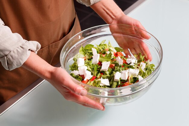 Foto vrouwelijke handen met een glazen kom groentesalade met feta over een glazen tafel