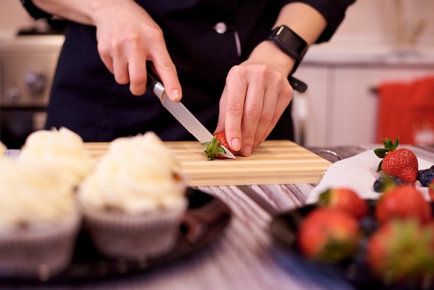 Vrouwelijke handen met een mes gesneden aardbeien op snijplank op de keukentafel. koken cupcakes met aardbeien en bosbessen.