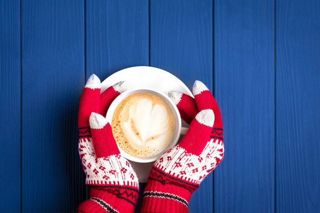 vrouwelijke handen met gebreide handschoenen met New Year's patroon houden witte kop met warme koffie cappuccino op blauwe houten