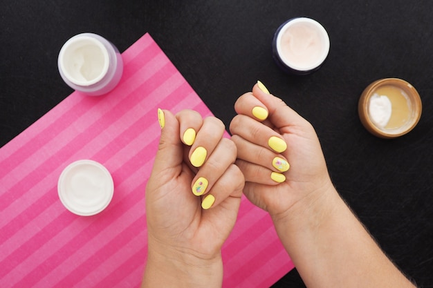 Vrouwelijke handen met heldere gele manicure. op de zwarte tafel staan potjes crème, vernis en een roze servet.