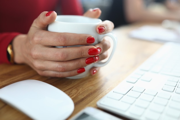 Vrouwelijke handen met rode manicure houden witte kop op werktafel