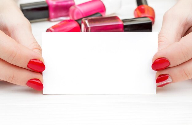 Vrouwelijke handen met rode manicure met visitekaartje en flessen met nagellak op witte houten tafel