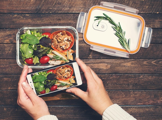 Vrouwelijke handen nemen foto van salade en kip lunchbox met smartphone