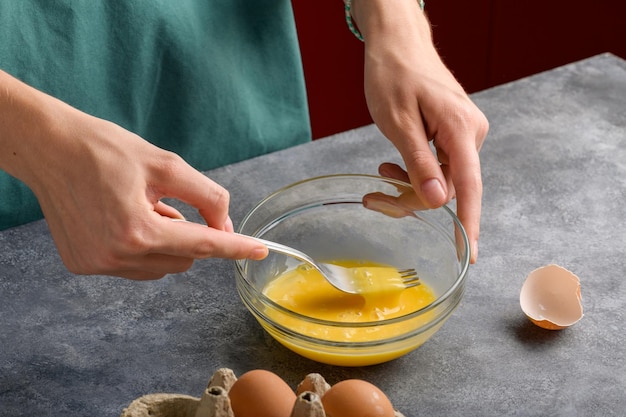 Vrouwelijke handen slaan eieren met een vork in een glazen schaal op een grijze keukentafel