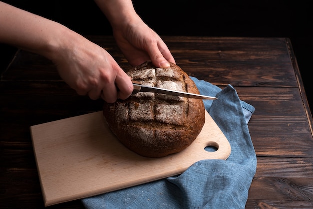 Vrouwelijke handen snijden een rond brood van roggebrood op een bord