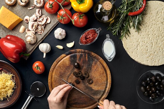 Vrouwelijke handgesneden olijven op een houten bord op de keukentafel rond leugeningrediënten voor pizza, groenten, kaas en specerijen