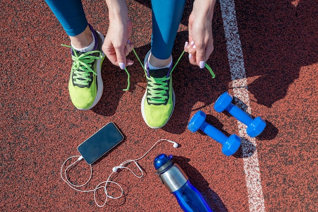 Vrouwelijke hardloper die haar schoenveters vastbindt en zich klaarmaakt om te racen op een atletiekbaan van een stadion met een fles water en een telefoon in een koptelefoon Van bovenaf bekijken