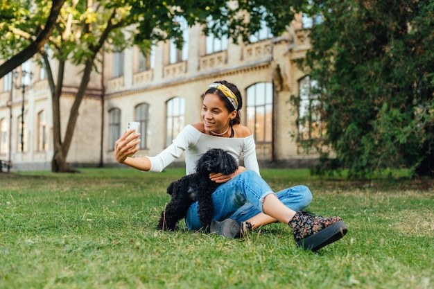 vrouwelijke hondeneigenaar zit op gras met huisdier en neemt selfie op smartphonecamera met glimlach
