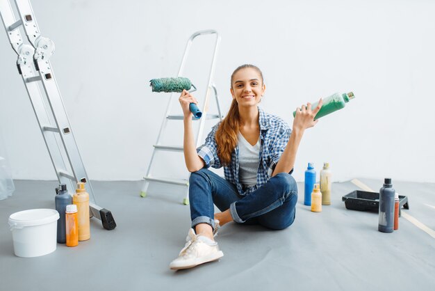 Vrouwelijke huisschilder met verf en roller zittend op de vloer. Huisreparatie, gelukkige vrouw die appartementsrenovatie doet