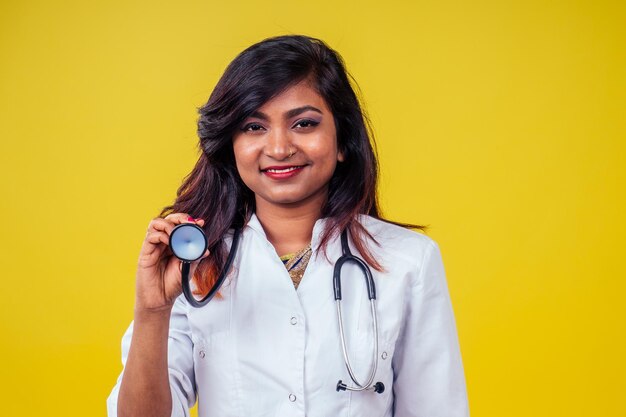 Vrouwelijke Indiase jonge en mooie blonde vrouw gynaecoloog arts met behulp van stethoscoop in een witte medische jas op een gele achtergrond in de studio.