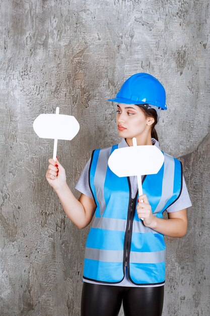 Vrouwelijke ingenieur in blauw uniform en helm met twee lege infoborden in beide handen.